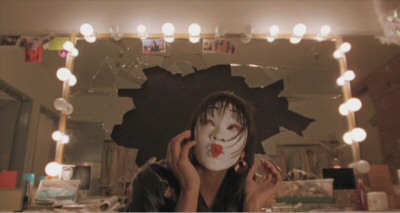 A girl in geisha makeup in front of a broken mirror, shielding her head.