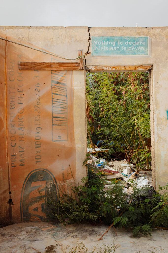 A photo from San Nicolas in Aruba, with a gaping abandoned doorway showing overgrown weeds behind it, and the words 