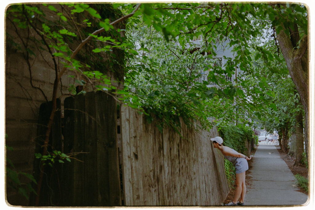Kat at a distance, leaning down to peer through a hole in a fence.