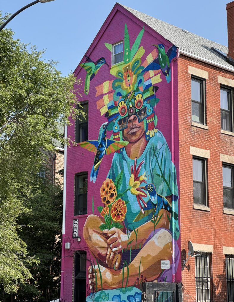 Street art of a young person with a colorful headdress, holding flowers and surrounded by hummingbirds