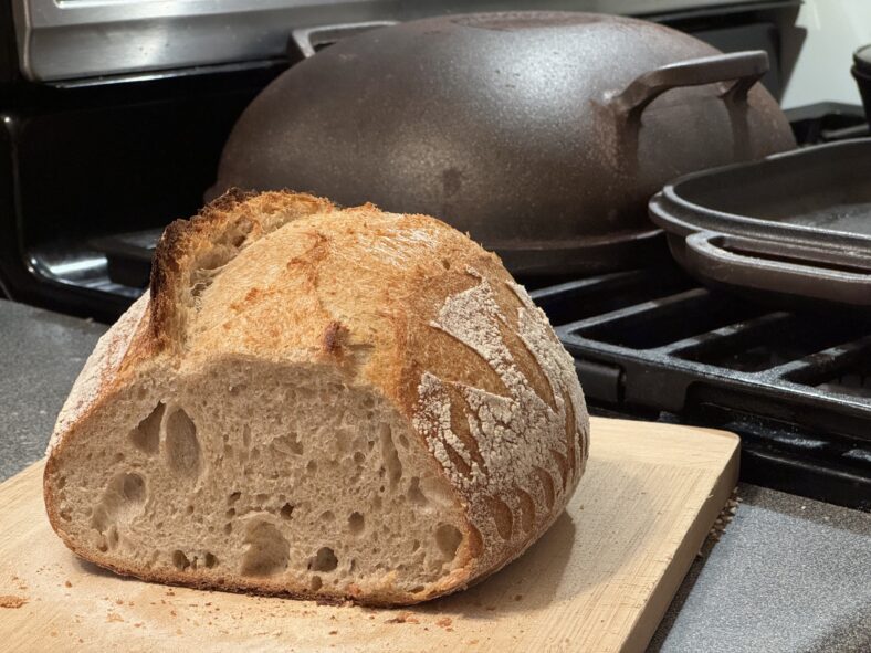 A small sourdough boule with its cut face toward the camera, and a cast-iron bread oven in the background