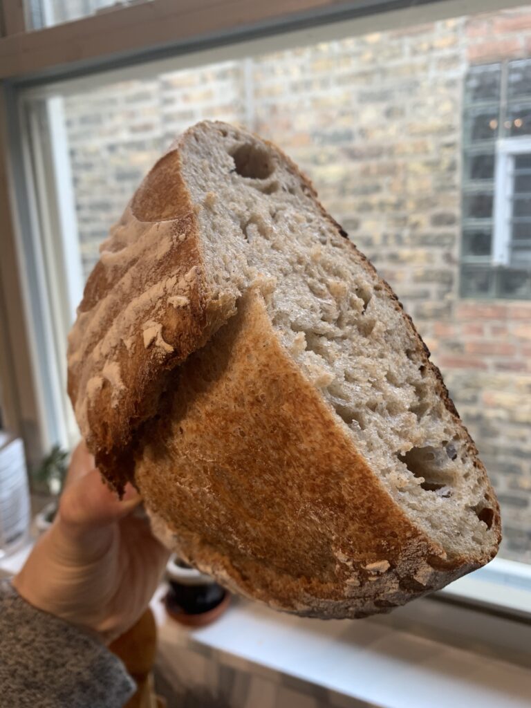 Half a loaf of sourdough bread, held up by hand in front of a window showing a brick wall opposite.
